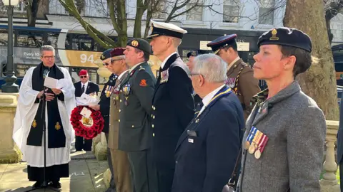 A rector reads a sermon at a war parade, as men and women in military uniform stand and listen. A double decker bus can be seen passing behind them in a town centre and there are trees and shops lining the road. It is a sunny day.