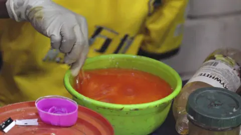 A seller wearing white rubber gloves in a market in Canon, mixes a substance orange in a green plastic bowl to create a product of the skin.