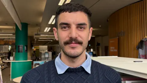 Image shows a man with a moustache and he is wearing a dark blue patterned jumper , underneath a light blue shirt collar is showing. He is stood infront of bookshelves in a library setting. 