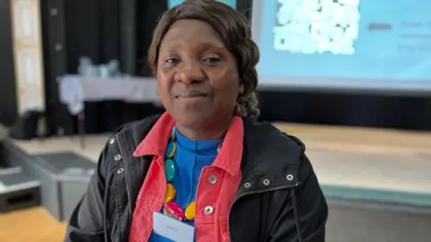 Nneka Iroegbu at Seaton Delaval arts centre has long brown wavy hair tied back. She is wearing an electrict blue top, with a coral denim jacket and a black coat. She is wearing a large colourful necklace and a name tag. She is slightly smiling at the camera.