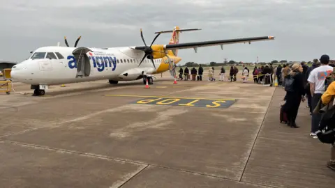 The image shows an Aurigny ATR aircraft with a queue of passengers snaking towards the plane. The aircraft has two propellers. Its tail is painted yellow while the rest of the fusillage is white. The plane has the word Aurigny on the side but the baggage door is open so the U is not visible. 