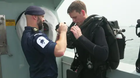 A picture of two people stood on a boat. The person on the right is wearing an all black diving suit.