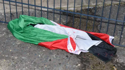 CPS A green, white red and black Palestine flag with flecks of red paint on it bundled up on the ground next to a blue fence
