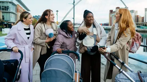 SolStock/GettyImages A group of women standing beside three prams on a riverside walkway in an urban area. They are dressed in winter coats and appear to be talking together. One person is holding an infant in a carrier on their chest, while another carries a baby wrapped in a blanket. Buildings, water, and a modern arched bridge are visible in the background.