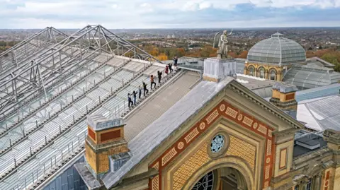 Wire & Sky People wearing safety harnesses walk along a metal walkway on the roof of Alexandra Palace, with the Angel of Plenty statue in the foreground and rooftops and trees stretching across north London in the distance under a cloudy sky.
