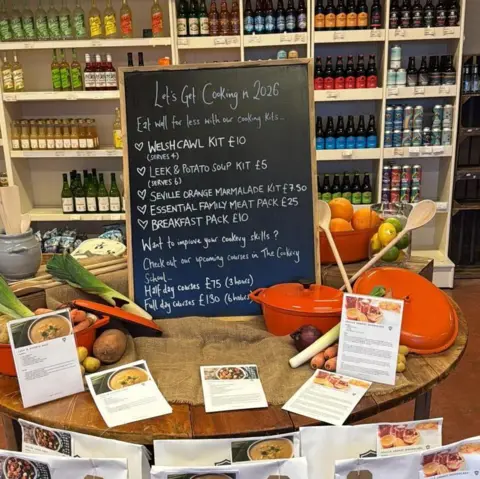 Olivia Parsons - Bodnant Welsh Food A chalkboard sign listing different Welsh dishes, such as Welsh cawl. In front of the sign is different vegetables. 