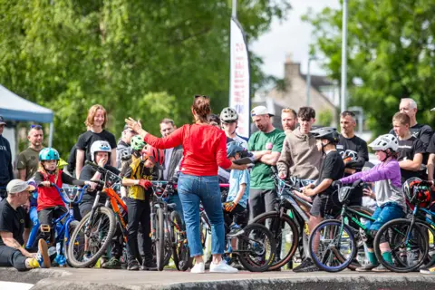 Pete Scullion A crowd of children on bikes and parents stands behind a woman in a red top with jeans who is explaining something to them