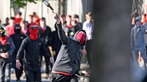 A young person with his face covered in a red mask throws a glass bottle of beer in the air towards a crowd behind the camera. Behind the young person is another person wearing a red balaclava and sunglasses. There are a crowd of people in the distance but their faces are blurred.