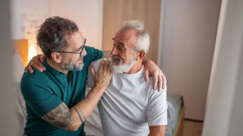 Getty Images Two men sat on a bed. The younger has an arm round the older man's shoulders and is wearing a green shirt and has visible tattoos on his arm