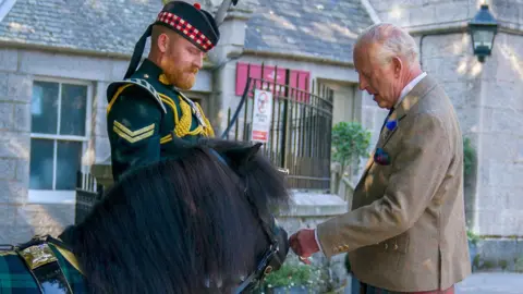 King Charles III - who is wearing tweed jacket, matching waistcoat, white shirt and blue tie; kilt, black shocks and black shoes - meets Shetland pony Cpl Cruachan IV (mascot of the Royal Regiment of Scotland) during an inspection of the Balaklava Company, 5th Battalion, The Royal Regiment of Scotland, at the gates of Balmoral, as he takes up summer residence at the castle.