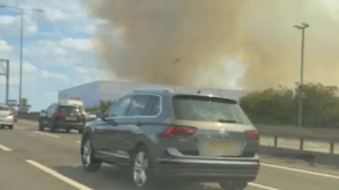 A silver family car is moving on a motorway. Other cars are in the frame, and in the background, large plumes of smoke rise from an industrial warehouse building.