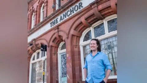 A man with brown hair and wearing a blue shirt is smiling and standing outside a pub with a sign above that reads "The Anchor".