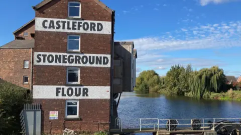 LDRS A brick building bearing the words 'Castleford Stoneground Flour' stands next to a river. On the other side of the river are several trees.