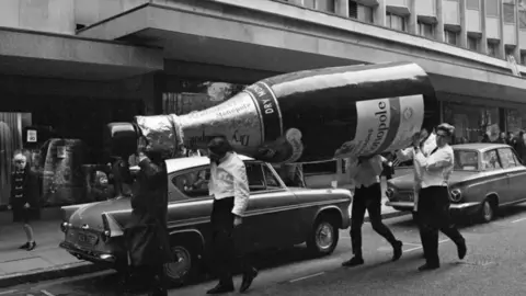 Getty Images A black and white photo of four people carrying a large champagne bottle waling into a department store. 