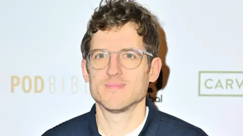 Getty Images Elis, who has brown curly hair, stubble and wears clear plastic-frame glasses. He is wearing a white t shirt and navy shirt and is looking at the camera with a neutral expression.