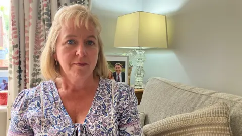 BBC Catherine O' Sullivan wearing a flowery blouse and sitting on her sofa, with a lamp in the background