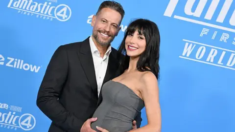 Getty Images Gruffudd standing with Wallace at The Fantastic Four: First Steps World Premiere. Gruffudd is standing on the left, wearing a black suit. He has one hand on Wallace's bump and the other hand is behind her back. Wallace wears a bandeau grey dress and has black straight hair. 