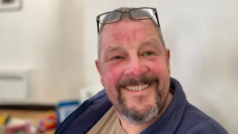 Mike Carson viste una chaqueta azul marino y una camisa beige. Tiene barba y sonríe. Sus gafas descansaban sobre su cabeza.