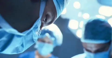 A surgeon wearing a mask and surgical hat in the foreground, with two other members of the surgical team wearing similar clothes in the background. All are looking down as they operate.