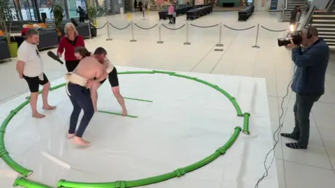 A cameraman films while two men wrestle each other in a green circle placed on a white sheet. They are bare chested and barefoot, with one wearing dark shorts and the other dark trousers. Next to them stands a man in a white tshirt and black shorts, talking to a woman holding a microphone. She is wearing a black top and trousers and a red jacket.