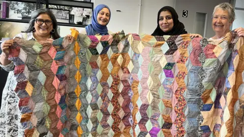 Four women smile, holding a colourful patchwork quilt.