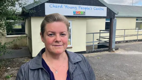 Serena Wootton looking at the camera. She is wearing a denim looking jacket over a blue top. A cream coloured single storey building behind has a large sign that says Chard Young People's Centre.