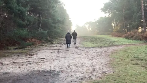 Luke Deal/BBC Two people walking in Sutton Heath woods away from the camera. They are wearing coats. There are trees lining the footpath either side. It looks like a frosty but sunny winter's day.