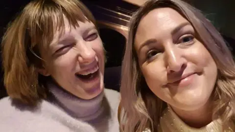 Two women are pictured in a selfie shot taken by the woman on the right. She has shoulder length curly hair and is smiling, her sister has shorter chin length hair with a fringe and is smiling widely