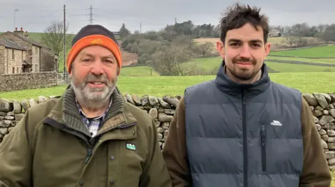 Two men stand in a field in front a dry stone wall. Beyond that a farm house, rolling hills and power lines.
