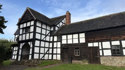 A two storey old farmhouse with white render and black wooden beams throughout the design.
