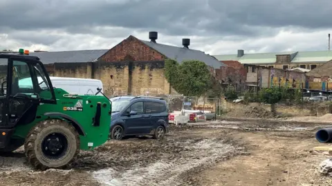LDRS A plot of apparent wasteland. A tractor and a 4x4 sit nearby. Some ramshackle old buildings are in the background.