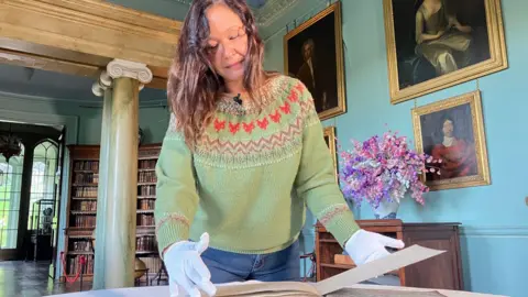 Coco Brown is wearing gloves and turning the page of the Red Book. She is standing in a grand room which has ornate marble columns, a full bookshelf, a bright flower arrangement and historic portraits are hung on the walls.