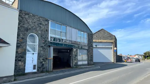 Two large former tram sheds made out of granite. Both have large hanger-style doors. In front a road and cycle path.