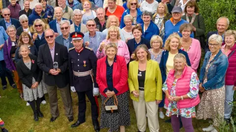A birds-eye view of a crowd, including a formally dressed Lord-Lieutenant looking up at the camera smiling. One person in the front holds a glass award.