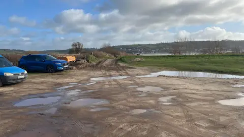 A car park with two blue vehicles parked. The ground is full of pot holes which are full of water and the beach is visible in the background.