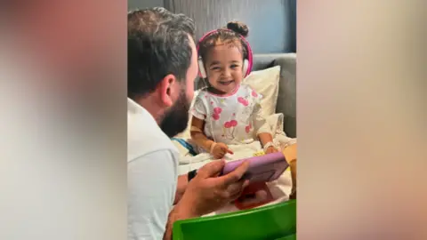 Family Aria is sitting on a bed smiling. She's wearing pink headphones. A man is holding a pink tablet pointing towards her.