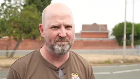 Un bărbat cu capul chel și barba încărunțită stă pe marginea drumului într-o zi înnorată.