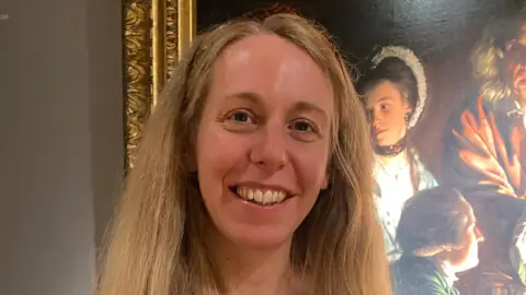 A woman smiles into the camera whilst standing in front of a painting which features dimly lit people.