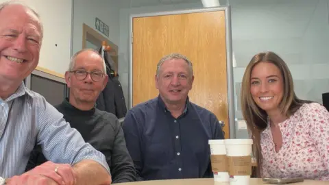 Supplied Prof John Wilson, Dr Mark Stoddart, Andrew Leslie and Bronwyn Mogie sit around a table and are smiling at the camera. John Wilson has short white hair and is wearing a blue striped shirt. Mark Stoddart is wearing a grey turtle neck and has short grey hair and glasses. Andrew Leslie has short greying hair and is wearing a dark blue shirt. Bronwyn Mogie has long brown hair and is wearing a pink top with a floral pattern on it.