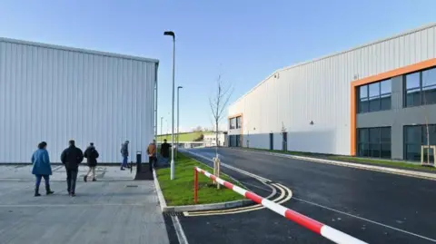 Two large white buildings with large windows. There are people walking behind one of the buildings on the left. There is a red and white stripped barrier on the right of the image, with a road at the centre, in-between the buildings.