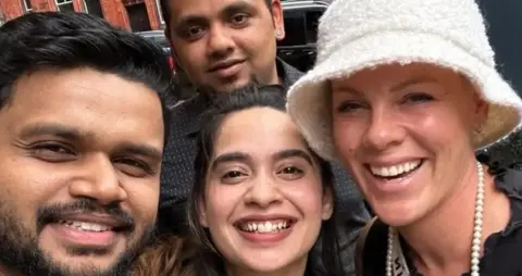 Asha's A close-up shows four people all looking at the camera with big smiles. Pink is on the right, and is where a white bucket hat and a white beaded necklace.