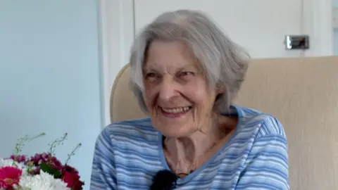A picture of Marjorie smiling at the camera. She is wearing a blue and white striped shirt with flowers next to her.