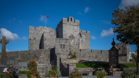 The exterior of Castle Rushen, a grey stone, small medieval castle with a clock tower, and a Manx flag on a clear day.