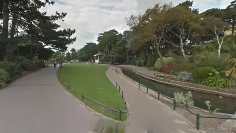 Grey paths either side of a green area and water stretching into the distance with trees and shrubs lining the sides