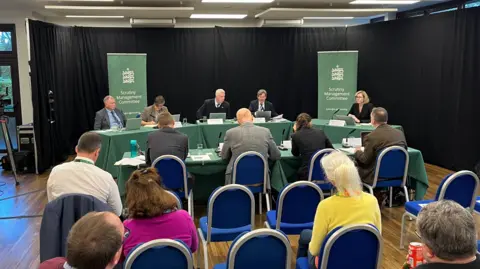 A panel of people sat behind a table, with a green cover. Behind them are two banners which state Scrutiny Managament Committee. In the audience are a few members of the public and some empty seats. 