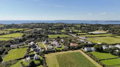 A drone shot of Sark on a bright blue sky day. It is green and lush and there are a number of mainly white houses surrounded by fields which have hedges as boundary markers. The sea can be seen and across the water, Guernsey.