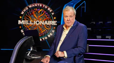 ITV Jeremy Clarkson looks at the camera from the Who Wants to be a Millionaire studio. He is sitting in the presenters seat wearing a blue suit.