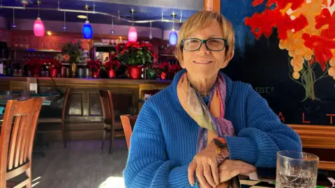 A woman wearing a blue sweater is stiing smiling in a colorful restaurant with a bar behind her with blue and pink lights over it. She is wearing a coloured scarf and there is a painting of red and yellow flowers behind her as well.