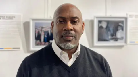 Rod Dixon standing in Bath Central Library. He is a black man with a bald head. He is wearing a black jumper with white shirt.