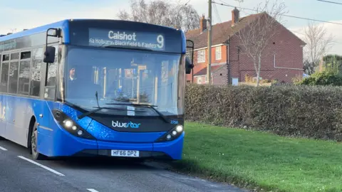 A Bluestar bus, which is a primary colour blue and has some white fleck detailing on the front, is driving along a residential road. It's information display at the front of the bus says it's the Calshot 9 service.